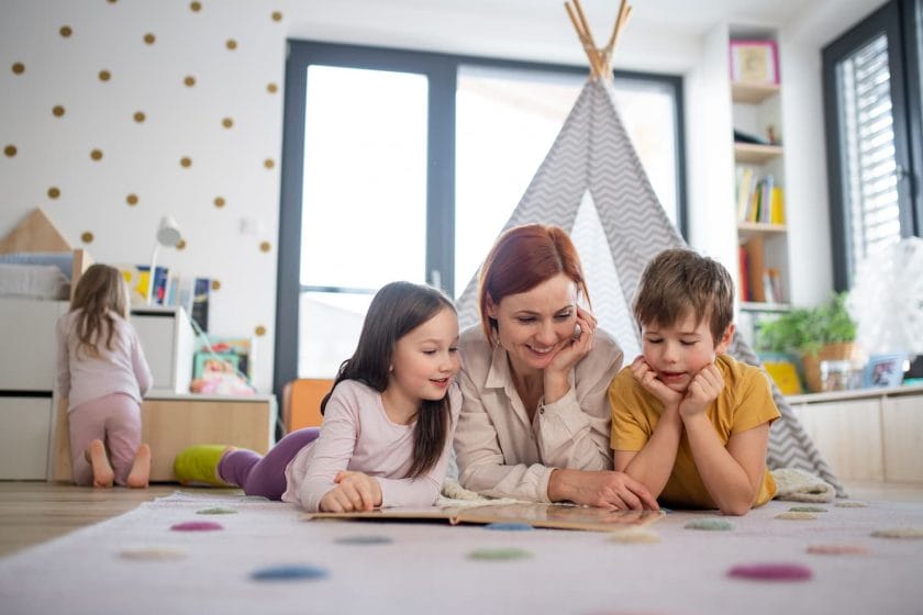 An image of a cheerful mother of three little children reading them book at home with a camping tent set up at the back.