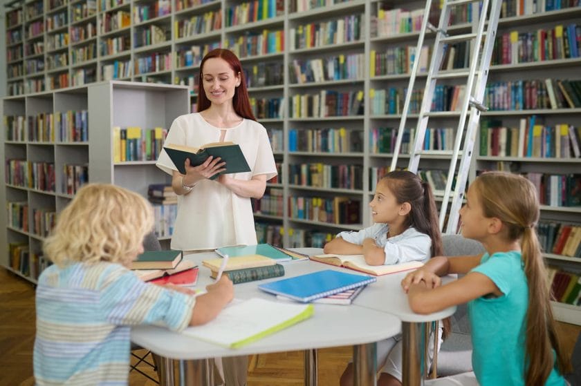 An image of a smiling woman with long red hair reading a book to children sitting at the table with textbooks listening with interest in the lightroom of the library.