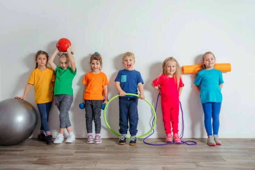 An image of a group of cheerful young kids in colorful and comfortable sportswear posing next to a white wall each holding one item of sports equipment.