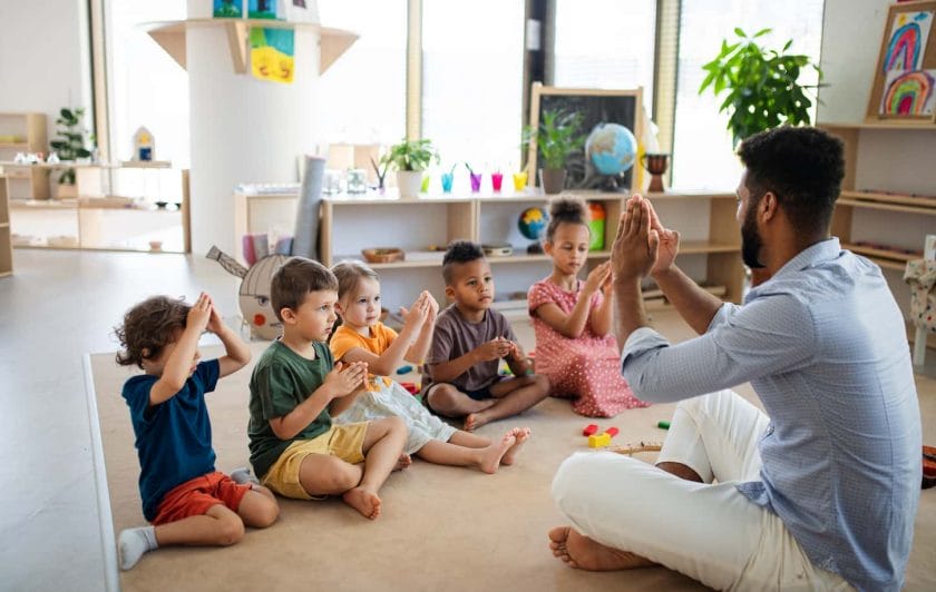 An image of Nursery school children with a man teacher sitting on the floor indoors in the classroom, playing.