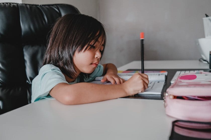 An image of a little girl writing in her textbook in an online class set-up.