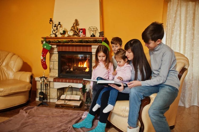 An image of a Happy young large family by a fireplace in a warm living room on a winter day. Mother with four kids at home read books.