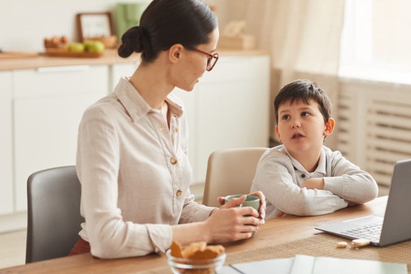 An image of a Warm-toned portrait of a young mother talking to her son while doing homework together sitting at the table in a cozy kitchen interior.