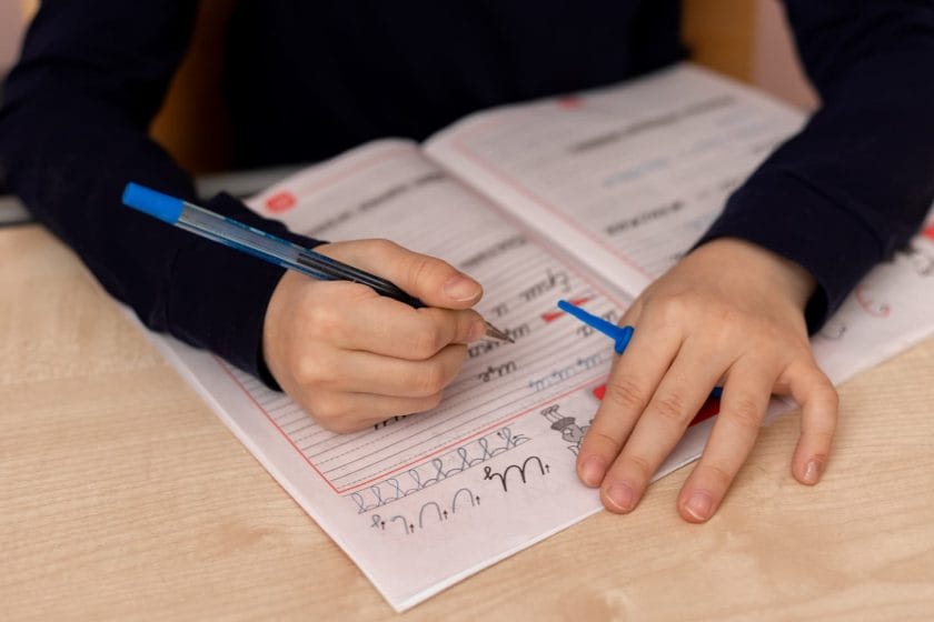 An image of A girl up to 10 years old doing homework on writing, learning to write blue capital letters with a pen.