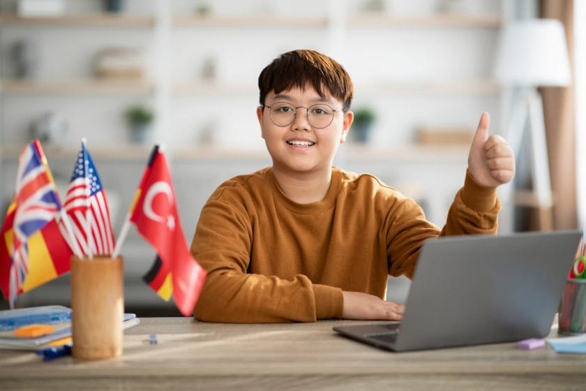An image of a Cheerful Chinese boy with a laptop and foreign flags are displayed on his desk.