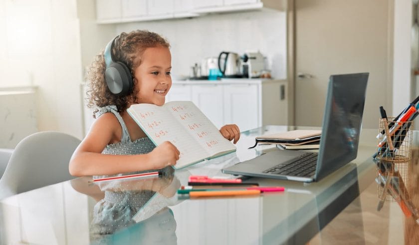 An image of Distance learning, education, and a little girl in a virtual on a laptop, smiling and showing her notes to her teacher.