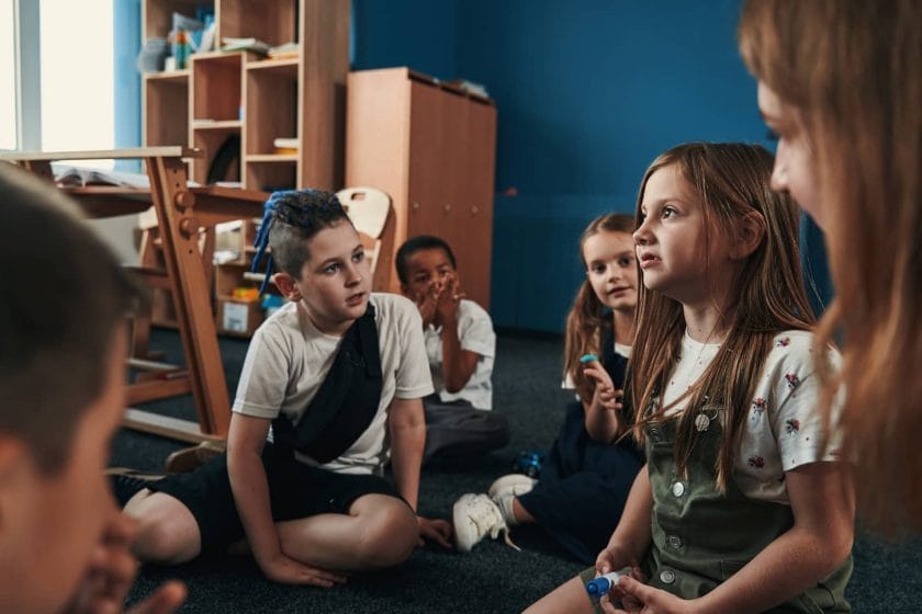 An image of a Thoughtful girl with a marker in her hands expressing her ideas while her classmates sitting on the floor around and listening.